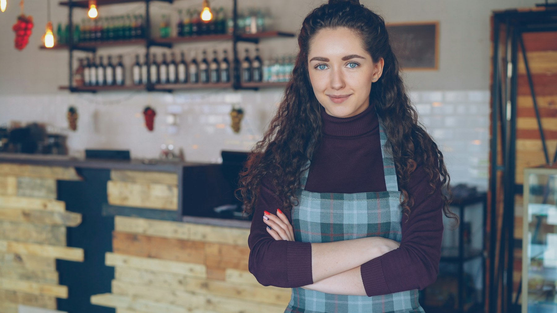 Lokales Café Eine lächelnde junge Frau mit Schürze steht mit verschränkten Armen in ihrem Café.