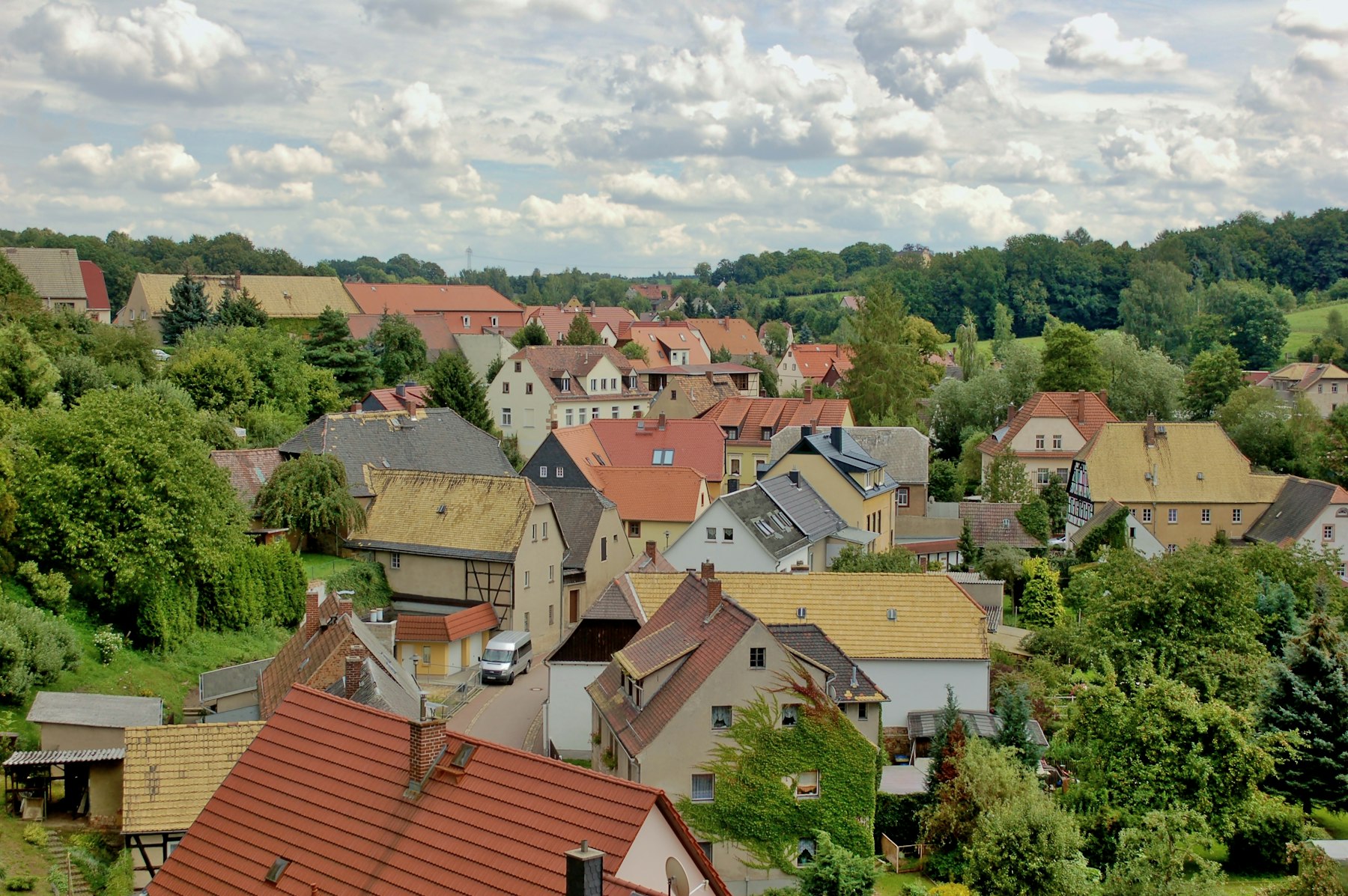 Blick über die Dächer eines Dorfes zwischen grünen Bäumen und Hügeln.