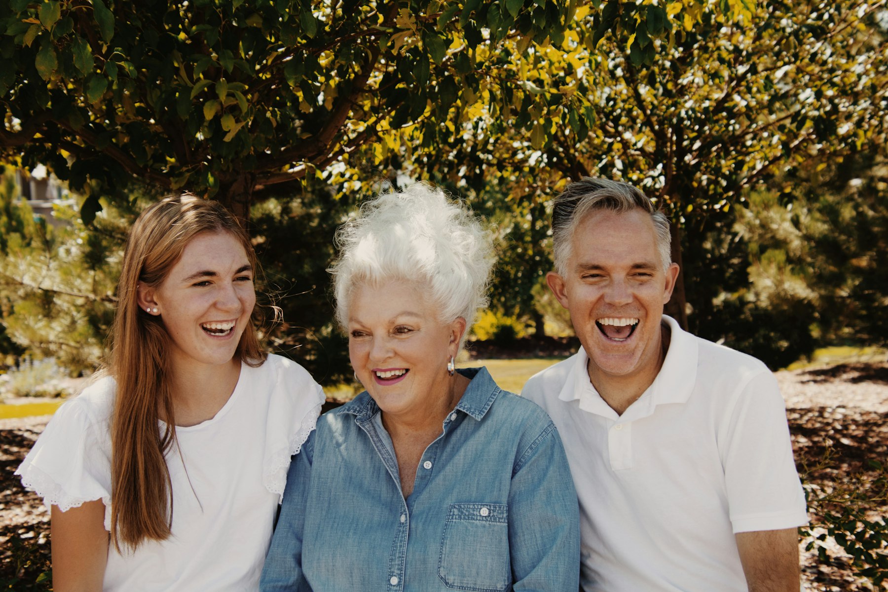 Drei Generationen lachen gemeinsam Eine junge Frau, eine ältere Frau und ein Mann sitzen im Grünen nebeneinander und lachen.